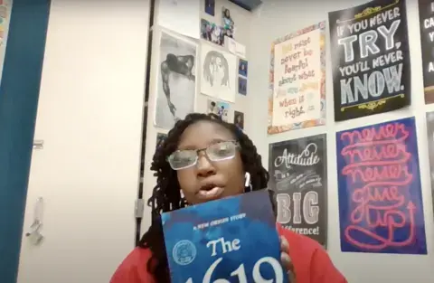 A young black woman wearing a red shirt and glasses holds a copy of the book "The 1619 Project: A New Origin"