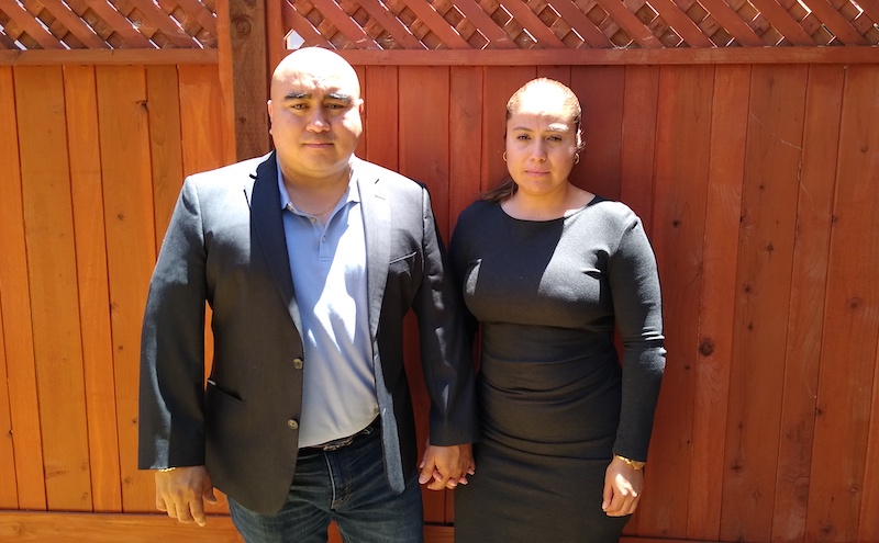 Jose Jaime & Gabriela Covarrubias, parents of Christian Madrigal, are standing together in front of a wooden fence. Jose Jaime & Gabriela Covarrubias, parents of Christian Madrigal, are standing together in front of a wooden fence.