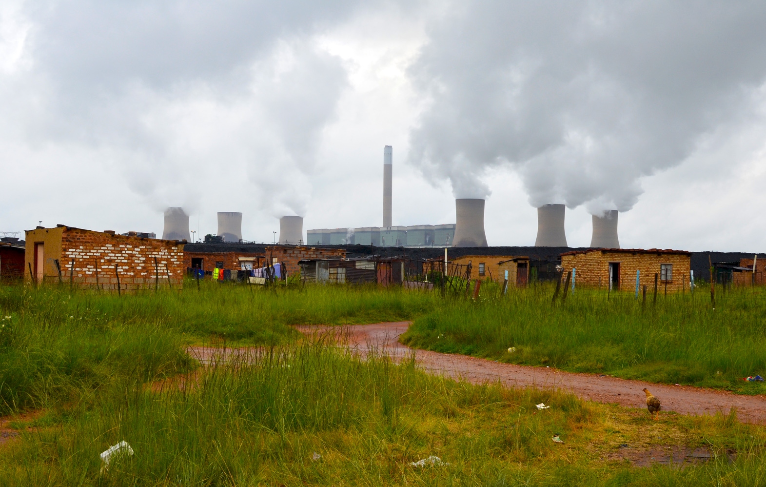 Duvha Power Station looms over the Masakhane informal settlement near eMalahleni in Mpumalanga. In 2015, the government pledged R18-billion to assist or relocate 15 mining-affected communities, including Masakhane. Image by Mark Olalde. South Africa, 2017. Duvha Power Station looms over the Masakhane informal settlement near eMalahleni in Mpumalanga. In 2015, the government pledged R18-billion to assist or relocate 15 mining-affected communities, including Masakhane. Image by Mark Olalde. South Africa, 2017.