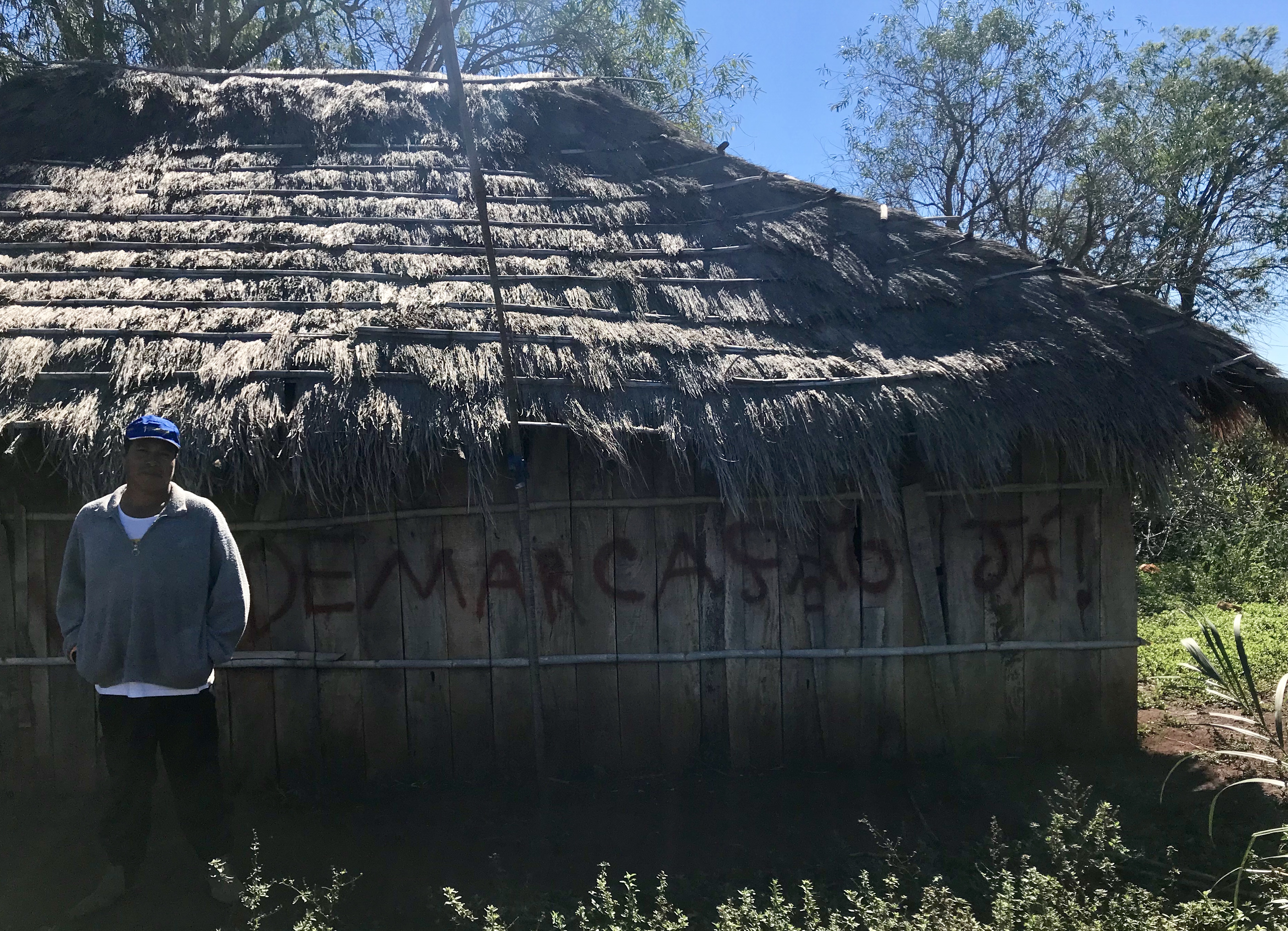 Cacique (or "Chief") Ezequiel João in front of the walls of his residence. "Demarcação Já!" (or "Demarcation Now!" is written as a form of protest to the government's long delay in demarcating and protecting the territories that were promised to his village. Image by Rafael Lima. Brazil, 2019. Cacique (or "Chief") Ezequiel João in front of the walls of his residence. "Demarcação Já!" (or "Demarcation Now!" is written as a form of protest to the government's long delay in demarcating and protecting the territories that were promised to his village. Image by Rafael Lima. Brazil, 2019.