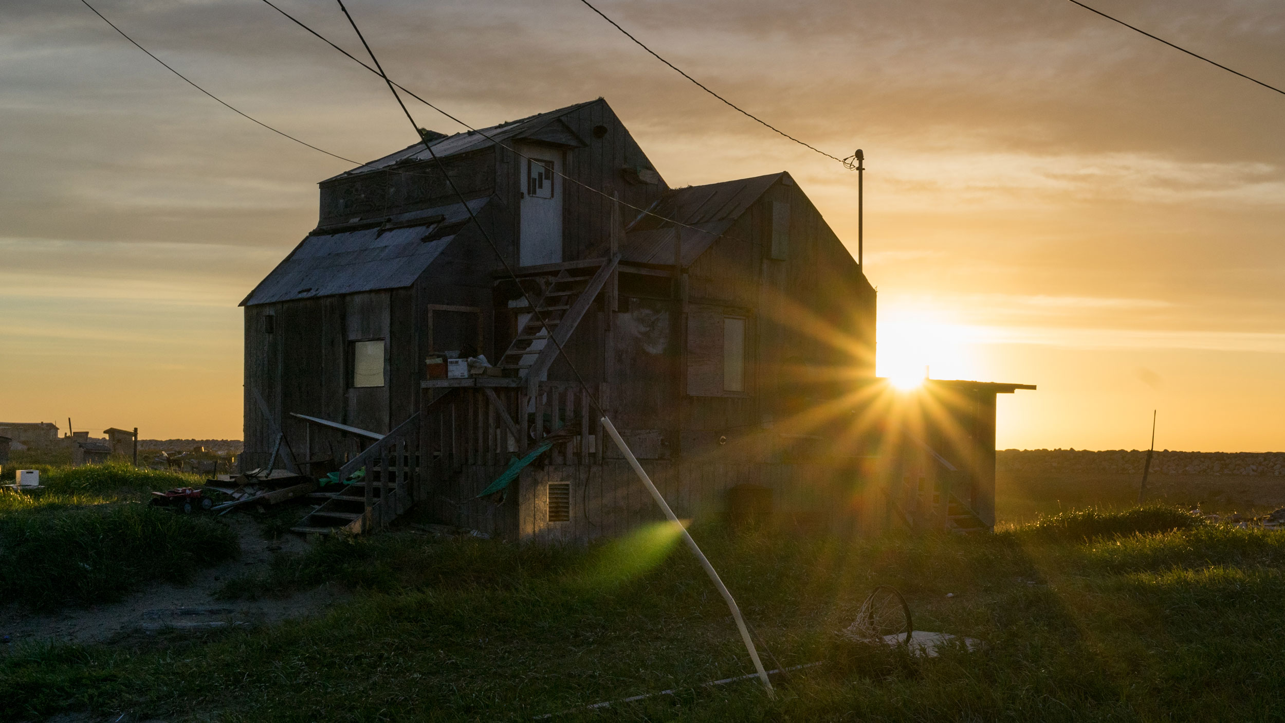 Rays of sunlight spill over a Shishmaref home as the midnight sun sets over the Chuckchi Sea. Image courtesy of Nick Mott. United States, 2018. Rays of sunlight spill over a Shishmaref home as the midnight sun sets over the Chuckchi Sea. Image courtesy of Nick Mott. United States, 2018.