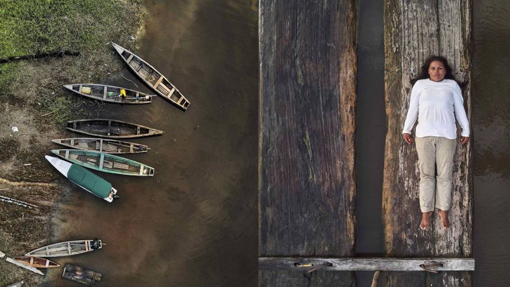 To the left, a series of empty wooden canoes stand by the margins of a river. On the right, Lilia Isolina Java Tapayuri lies down on a plank of wood, looking directly at the camera, captured from an aerial shot. She is wearing a white long sleeved shirt, khaki-colored pants, and no shoes.
