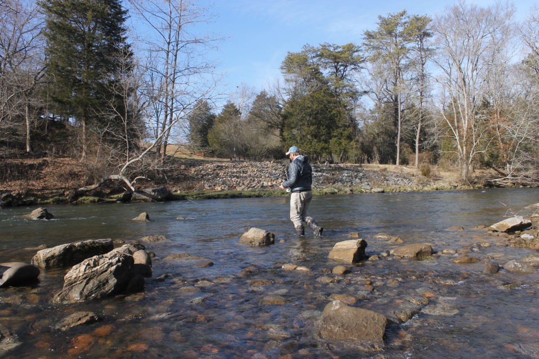 A man with a fishing pole wades through a river. The water is about just above his ankles.