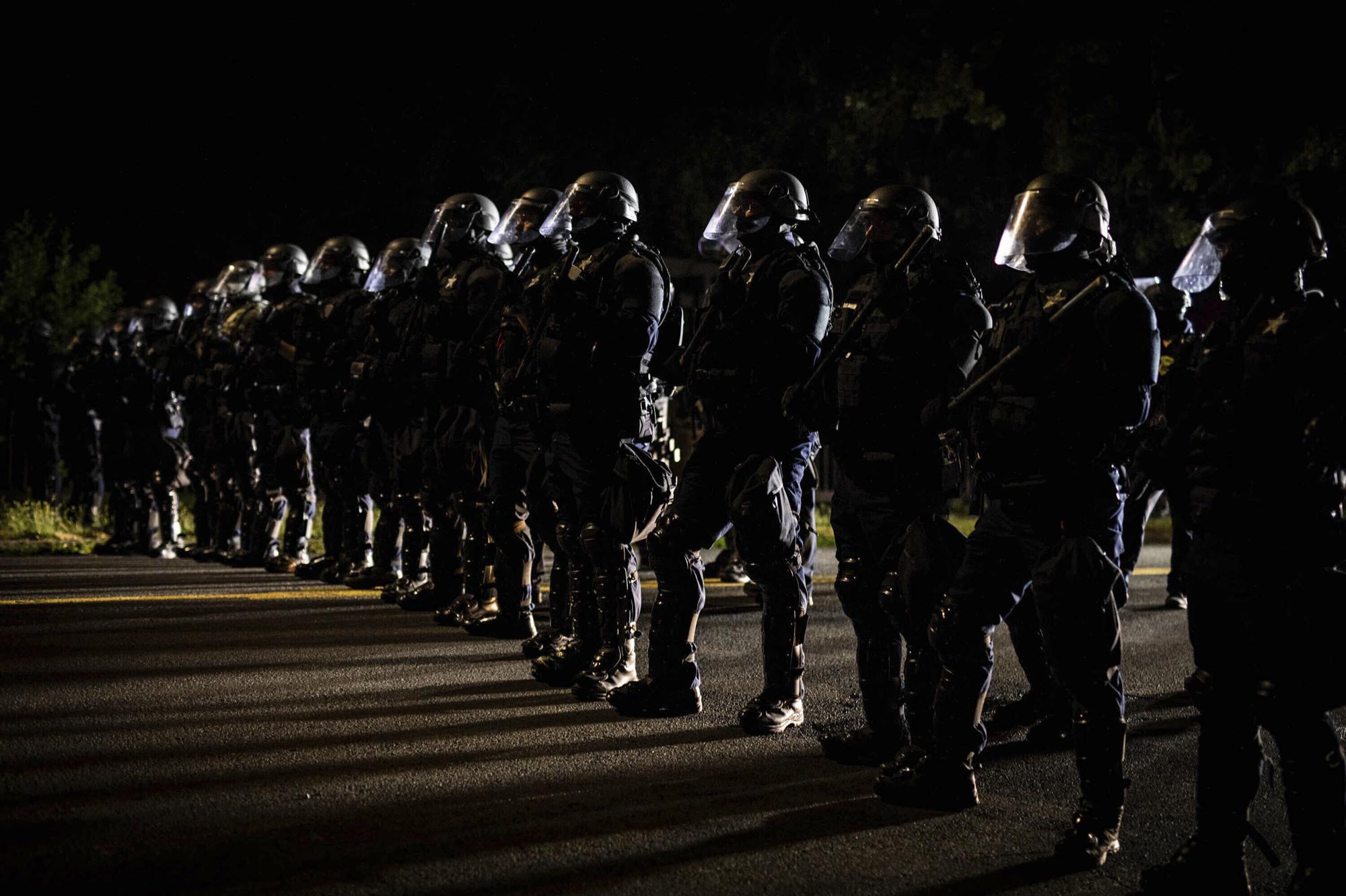 Police officers in riot gear line up across a street.