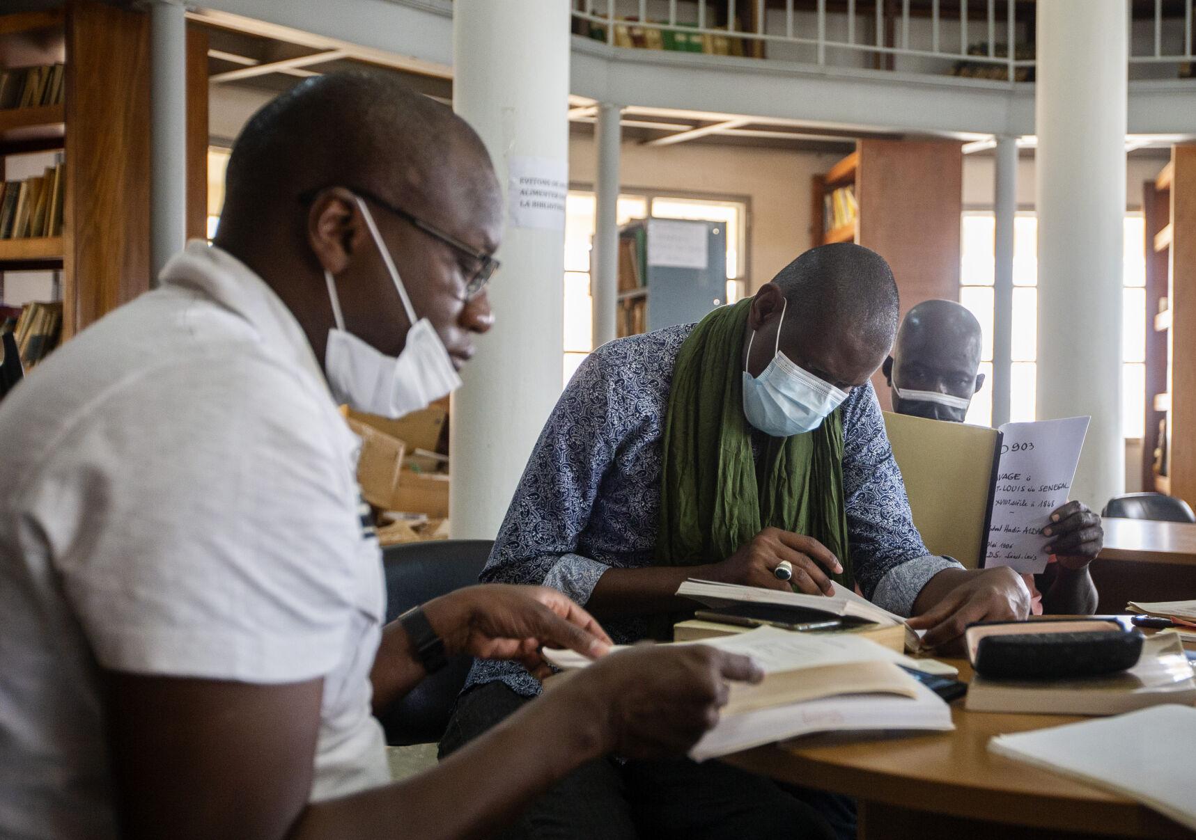 Three people wearing medical face masks sit and read papers and books in an archive.