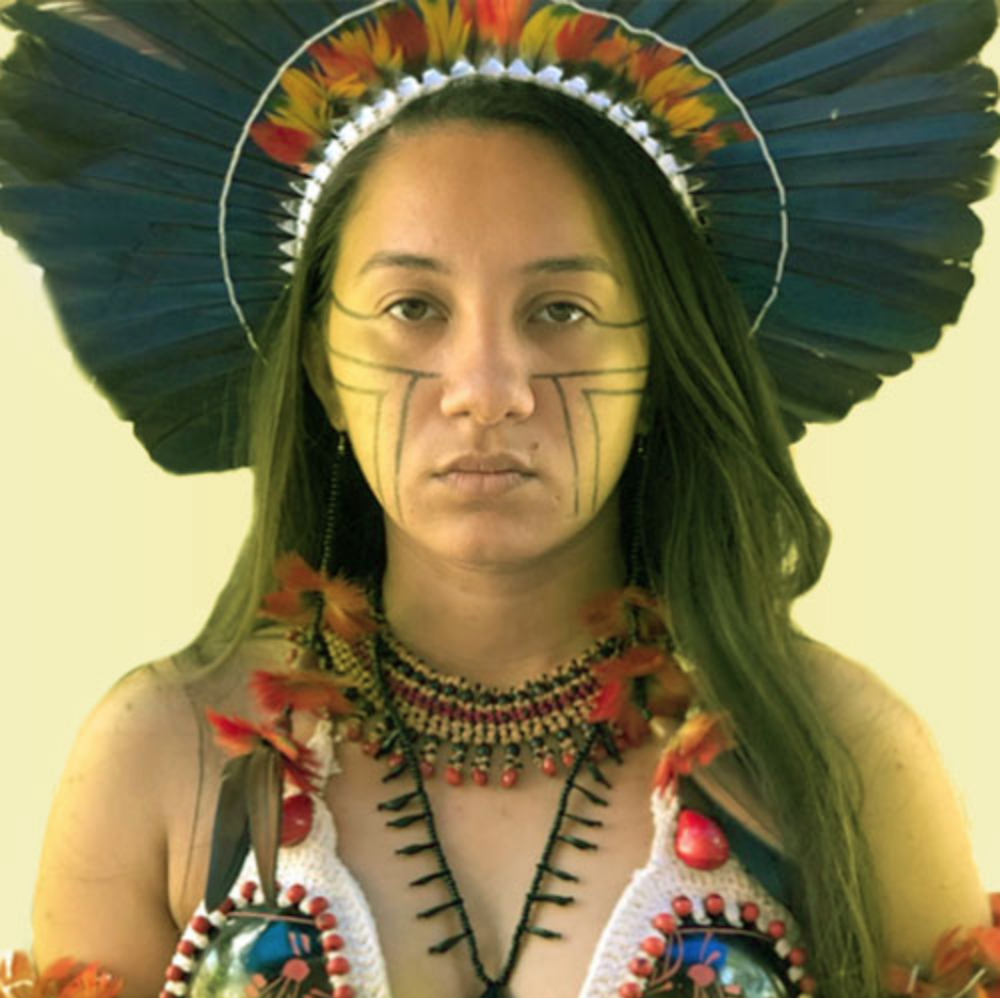 A person with black hair and Indigenous headdress looks into the camera.