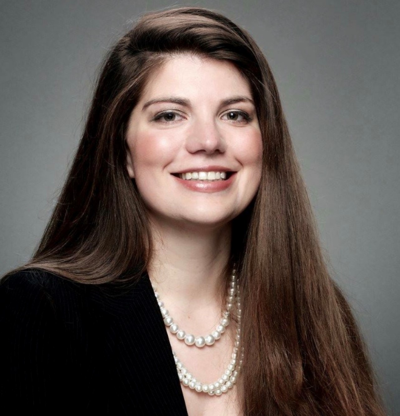 A woman with long dark hair wearing a black blazer against a grey background