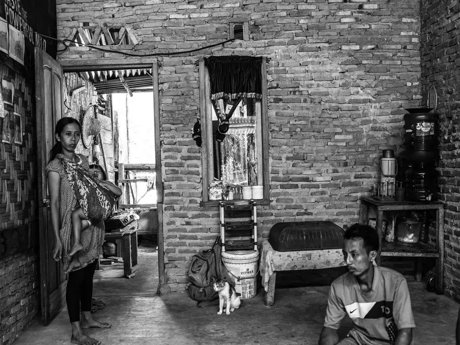 AFTER A DAY'S LABOR.&nbsp;Ida tries to comfort her daughter Santi while her husband Supriyono sits on the floor after a day's work. The gallon jugs of water in the corner are their only water for two weeks. They pay 56,000 Rupiah ($3.84) per gallon. Image by Xyza Cruz Bacani. Indonesia, 2018.