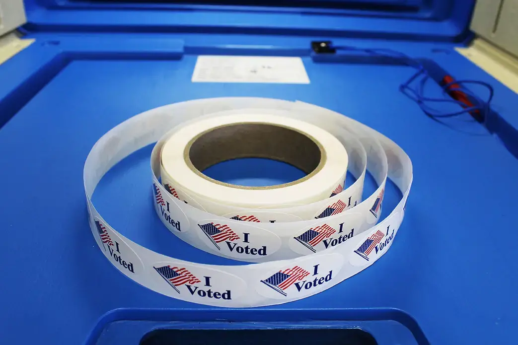 Red white and blue "I voted" stickers with American flag are on a spool of stickers placed on a voting booth platform. Image by Barbara Kalbfleisch. United States, undated. 
