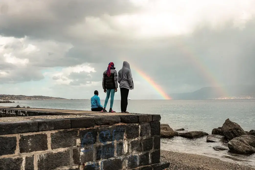 Blessing and two other teen-age Nigerian girls watch a rainbow over the short stretch of water separating Sicily from mainland Italy. Eighty percent of young Nigerian women who cross the Mediterranean are trafficked into sexual exploitation. Image by Ben Taub. Italy, 2017.