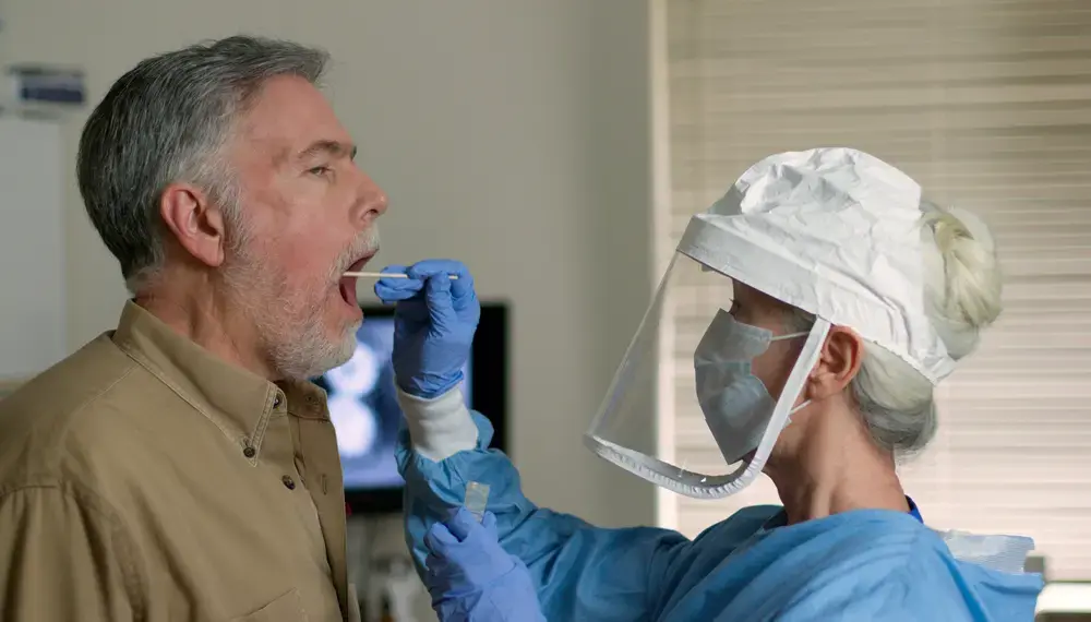 A man being swabbed by a healthcare worker in protective garb to determine if he has contracted the coronavirus. Image by JHDT Productions / Shutterstock. United States, 2020.