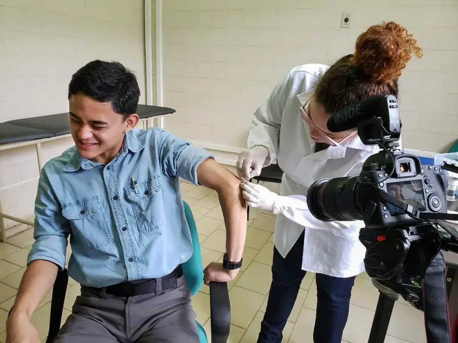 2020 Reporting Fellow Anton L. Delgado grimaces as Raquel Bouth, a Ph.D. student studying microbiology, takes a sample from the olecranal skin on his left elbow. This was the final part of Delgado’s leprosy evaluation that he took after nearly a month of field work in northern Brazil. Image by Moises Silva. Brazil, 2020.