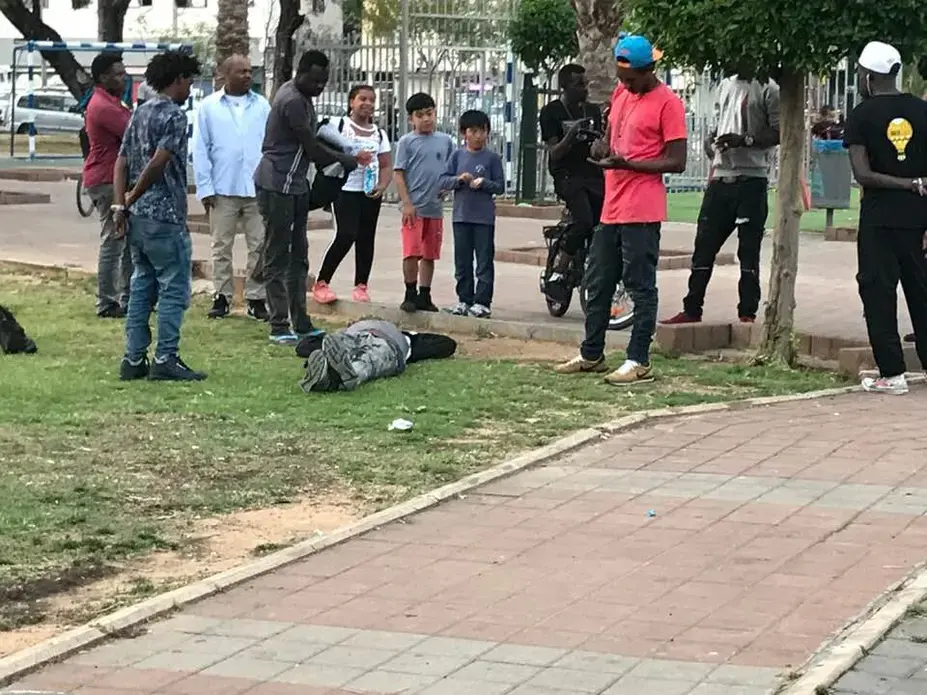A Sudanese refugee reacts to substance overdose and bystanders surround him in Levinsky Park in south Tel Aviv, Israel. Many refugees suffer various mental health disorders such as post-traumatic stress disorder (PTSD), anxiety, depression, alcohol and substance abuse, and in extreme cases, psychosis. Image by Isma'il Kushkush. Israel, 2018.