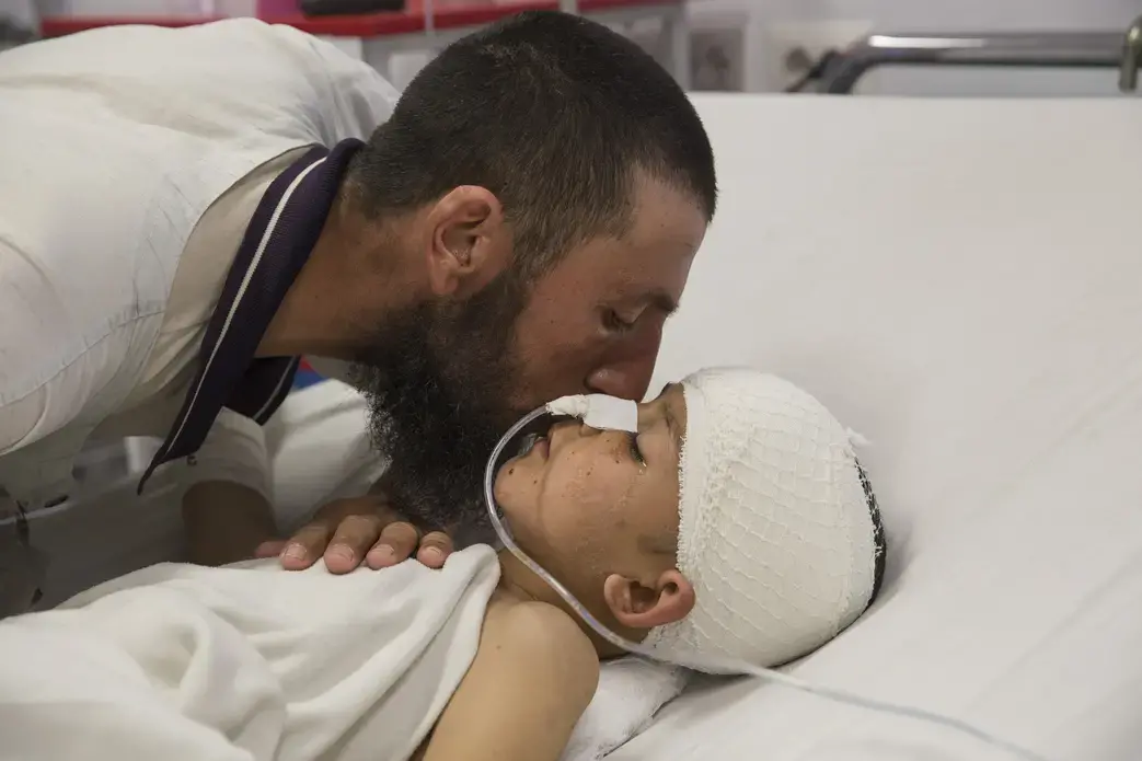 Abdul Kabir kisses his son, Noor Ahmad, 8, who is unconscious with a severe brain injury from an improvised explosive device (shell injury) from Mazar-E-Sharif Kabul on March 12, 2016. His father was also injured but recovered. Image by Paula Bronstein. Afghanistan, 2016.