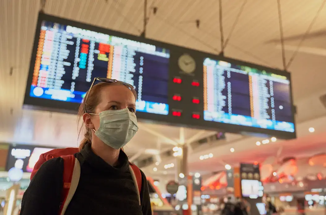 A woman with a backpack wearing an anti-virus mask on her face at the airport arrivals board. Image by Damian Pankowiec/Shutterstock. 2020.