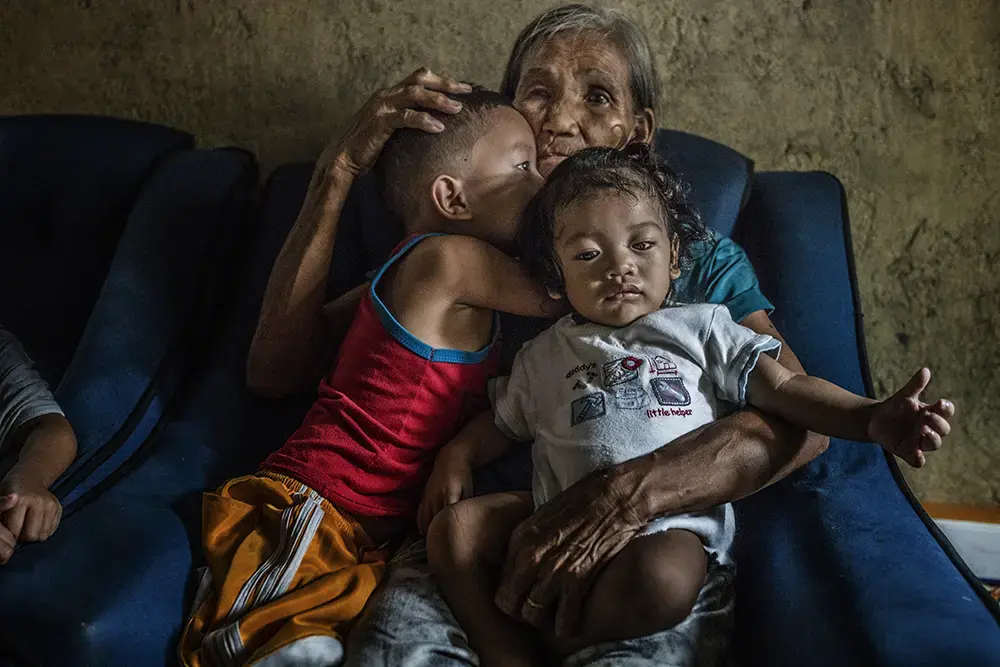 Remy Fernandez, 84 years old, holds two of the seven grandchildren she is raising. Her youngest son, Constantino de Juan, a methamphetamine user, was killed by masked men in December, 2016. Upon seeing his attackers, Juan instructed five-year-old CJ, shown here wearing a red tank top, to take care of his siblings. The children's mother is in prison due to a drug arrest. Baby RJ, in the "daddy's little helper" T-shirt, was born in prison. Image by James Whitlow Delano. Philippines, 2017.