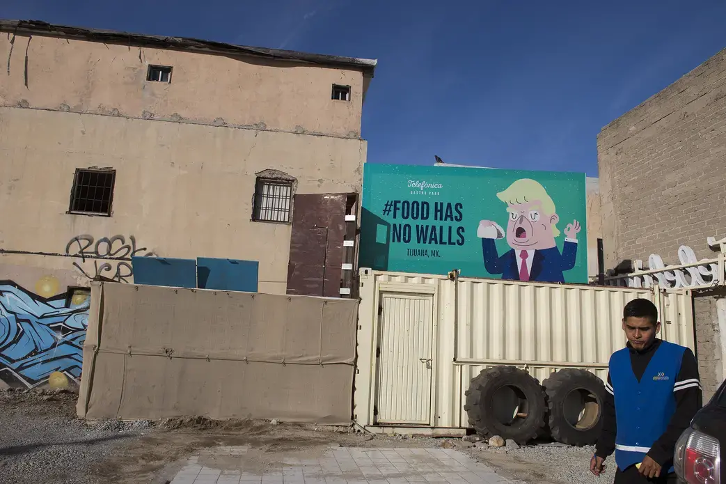 A billboard advertising Telefonica Gastro Park, an area in Tijuana featuring a food cart-like dining in Zona Centro, features artwork with the likeness of President Donald Trump as a parking valet works nearby Dec. 1. Image by Amanda Cowan. Mexico, 2019.