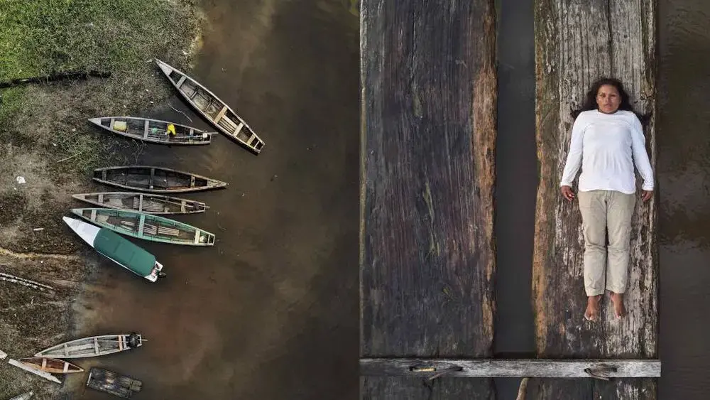 À esquerda, uma série de canoas de madeira vazias estão às margens de um rio. À direita, Lilia Isolina Java Tapayuri deita-se em uma prancha de madeira, olhando diretamente para a câmera, capturada em uma foto aérea. Ela está vestindo uma camisa branca de mangas compridas, calça cáqui e sem sapatos.