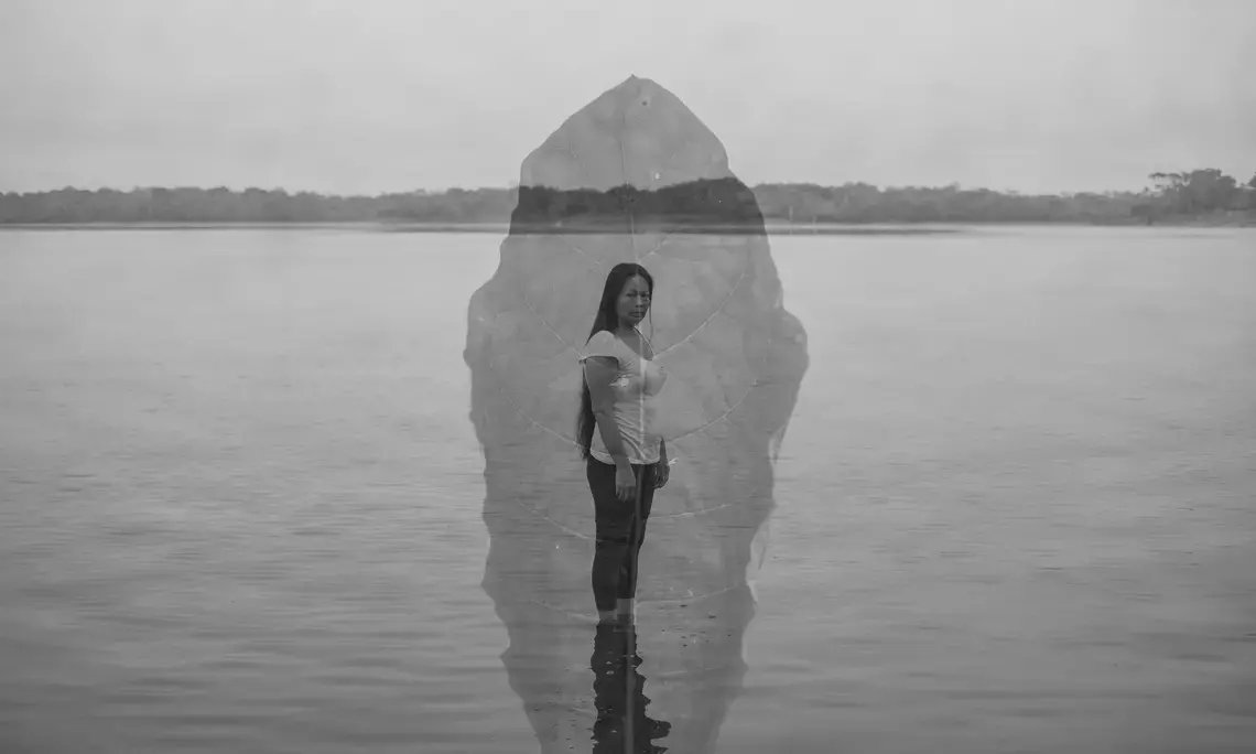 Black and white double exposure photo of a woman standing in front of a body of water with a leaf superimposed on the image.
