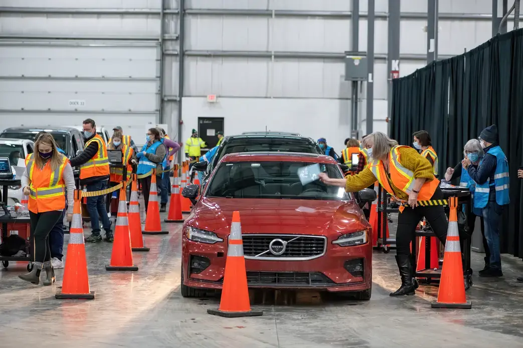 Workers in masks and reflective vests distribute vaccines as people wait in cars.