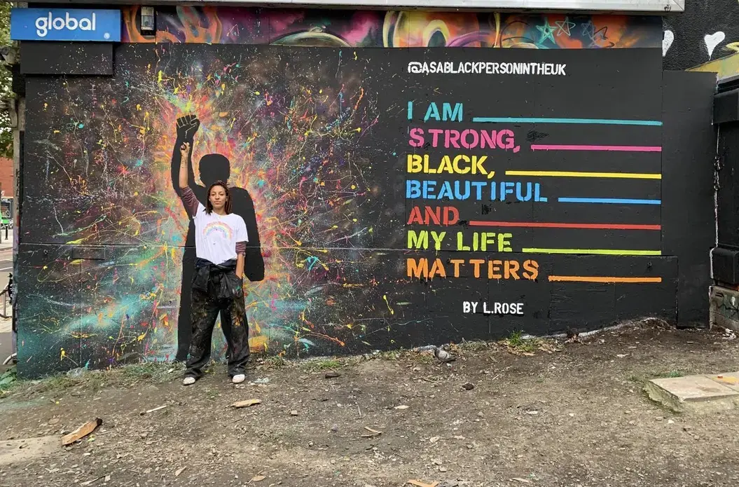 A woman stands in front of a mural with her fist raised. Her pose is echoed by a figure in the mural. Text on the wall reads, "I am strong, Black, beautiful, and my life matters."