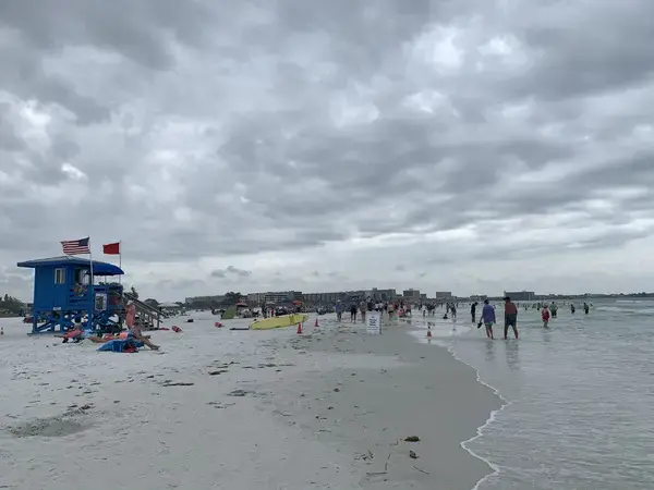Image of beach with a blue lifeguard tower on the left of the frame and the ocean on the right. Dozens of beachgoers sit on the sand or walk through the tide. A few carry surfboards.