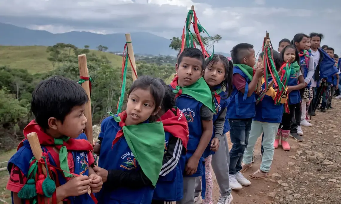 Children in blue vests and green-and-red bandanas tied around their necks line up on a mountain trail. Each carries a wood staff.