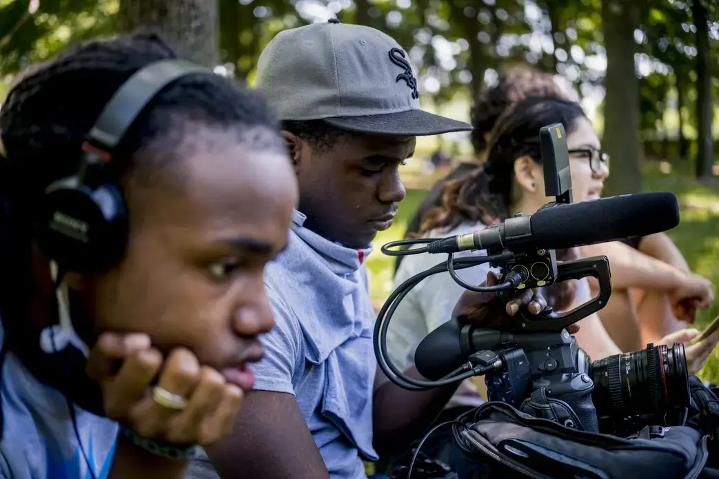 Four students work on creating a documentary. In the foreground, a student wears headphones to listen to audio they are recording. Another student uses a video camera to film. In the background, two students sit and watch the scene.