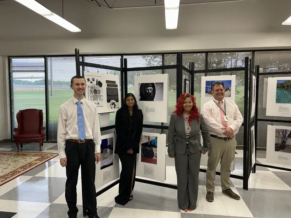 Three students and their teacher pose in front of the display of images from the How I See It Photography Exhibition