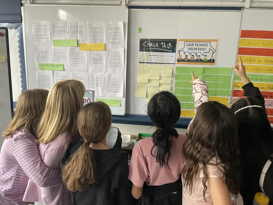 group of young girls face the whiteboard and point to documents taped to the board
