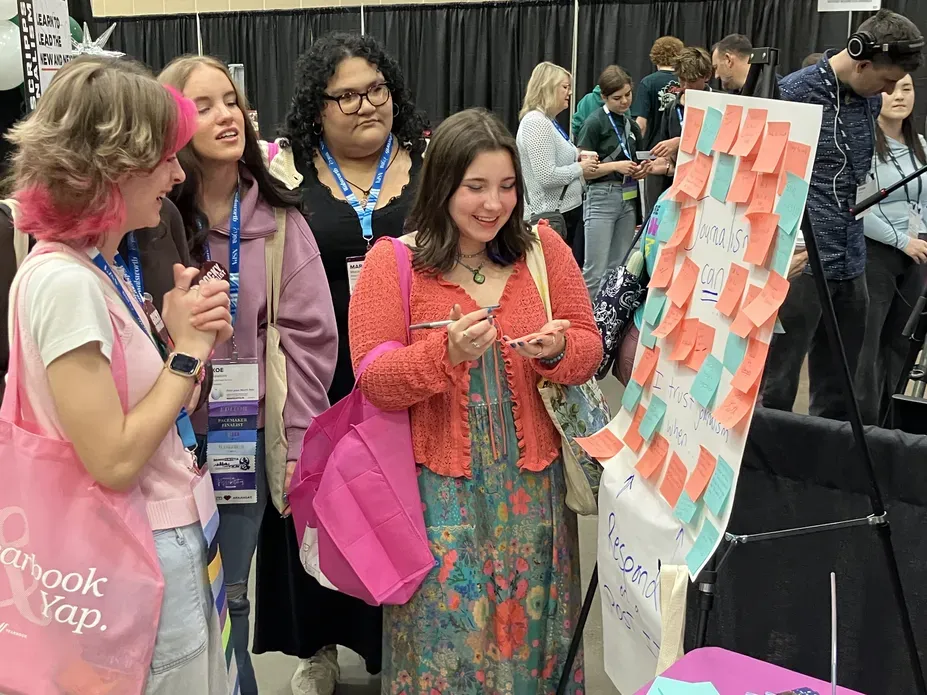 Three students and a teacher talk, laugh, and fill out post-its to stick on a posterboard in a conference exhibit hall.