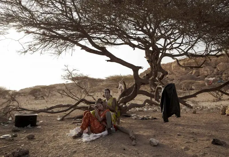 Ethiopian migrant Fatma, right, braids her friend's hair as they take shelter in Obock, Djibouti, at the last stop of their journey before leaving by boat for Yemen in the evening, July 15, 2019. Image by AP Photo / Nariman El-Mofty. Djibouti, 2019.