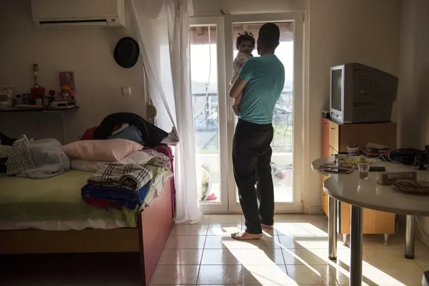 Yousuf Arsan, 27, sings to baby Rahaf, almost 6 months, while his wife (not shown) Noor Al Talaa, 22, cooks lunch at an apartment in Sidros. Image by Lynsey Addario. Greece, 2017.