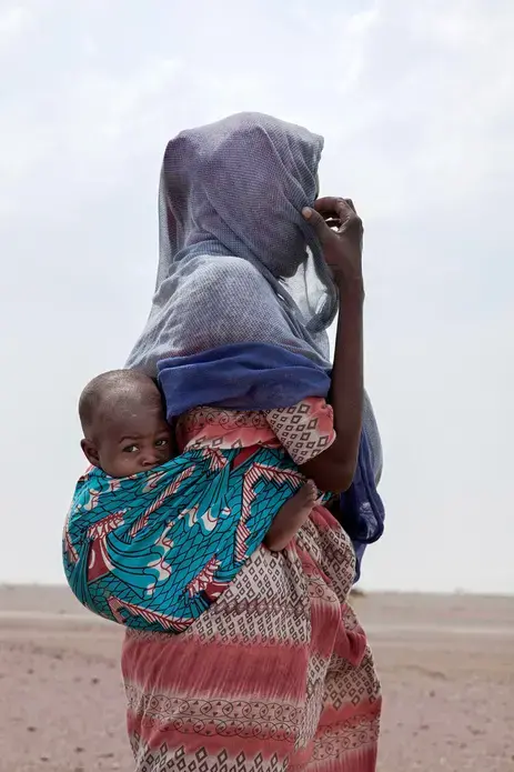 In this July 23, 2019 photo, Fatma, a migrant from Mali, carries her one-year-old Said Yaacoub, as she makes her way in Lahj, Yemen. Fatma and her family of four are slow because of the children. They came by boat from Djibouti and dream of going Saudi Arabia. They were taken to the lockups known in Arabic as 'hosh' but smugglers let them go because of the children. Image by Nariman El-Mofty / AP Photo. Yemen, 2019.