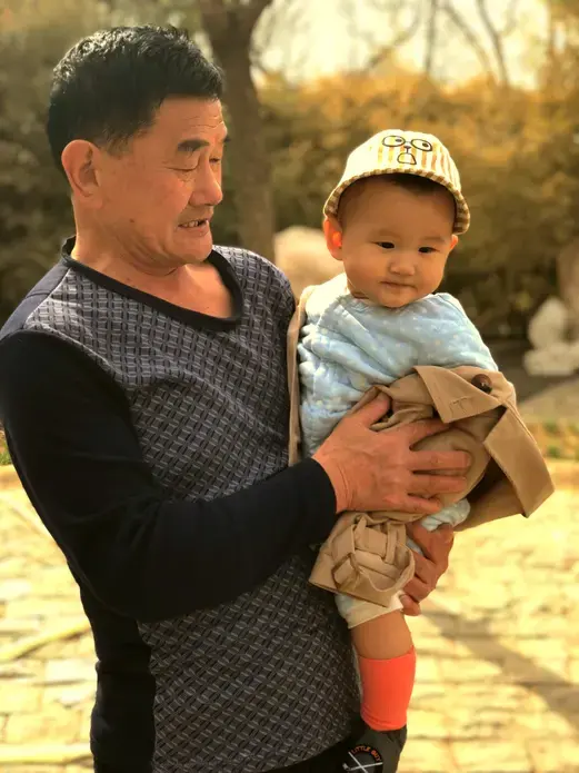 At a park in Beijing, an elderly woman rolls a stroller while this man carries a baby boy. Image by&nbsp;Argentina Maria-Vanderhorst. China, 2018.