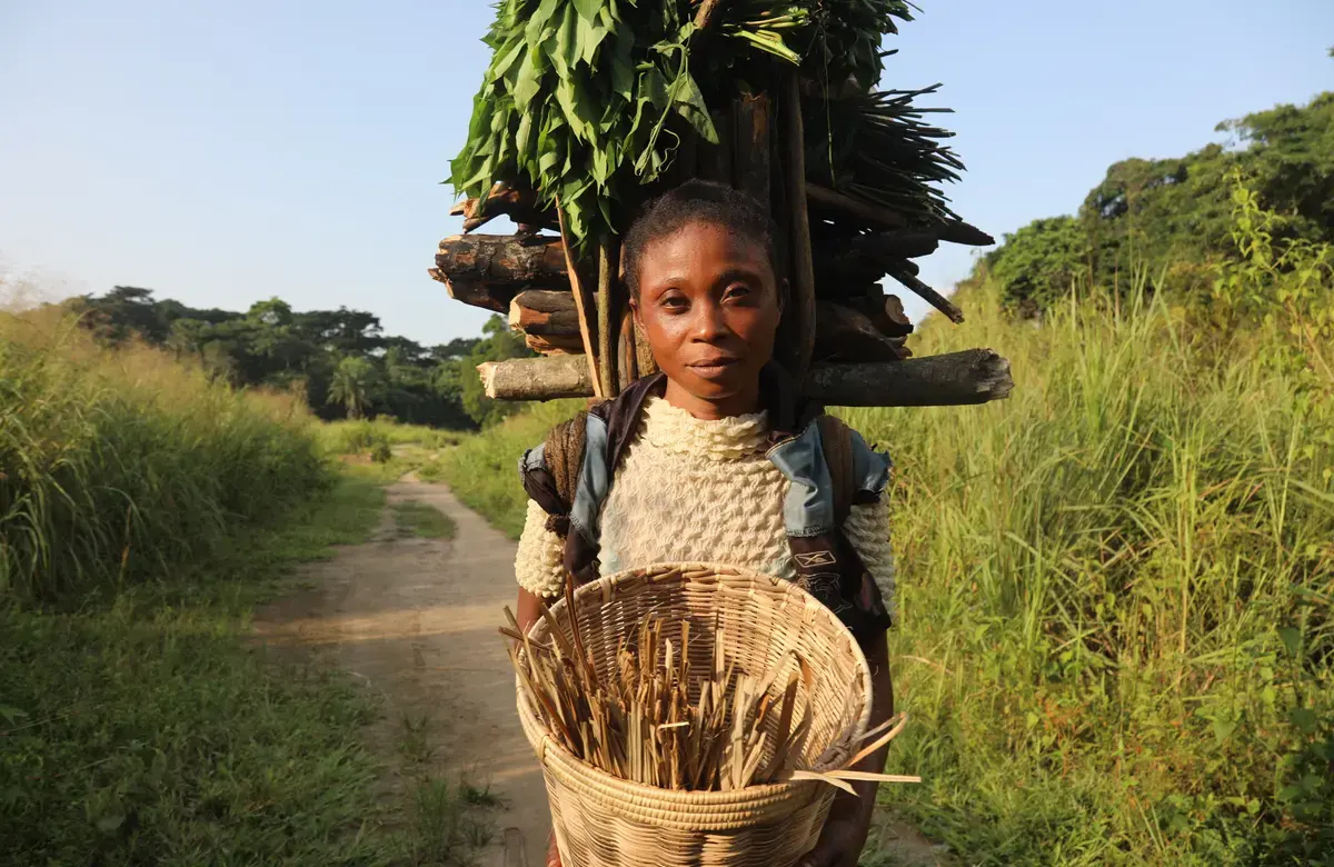 A woman faces the camera and holds an empty woven basket with more wood and plants tied to her back