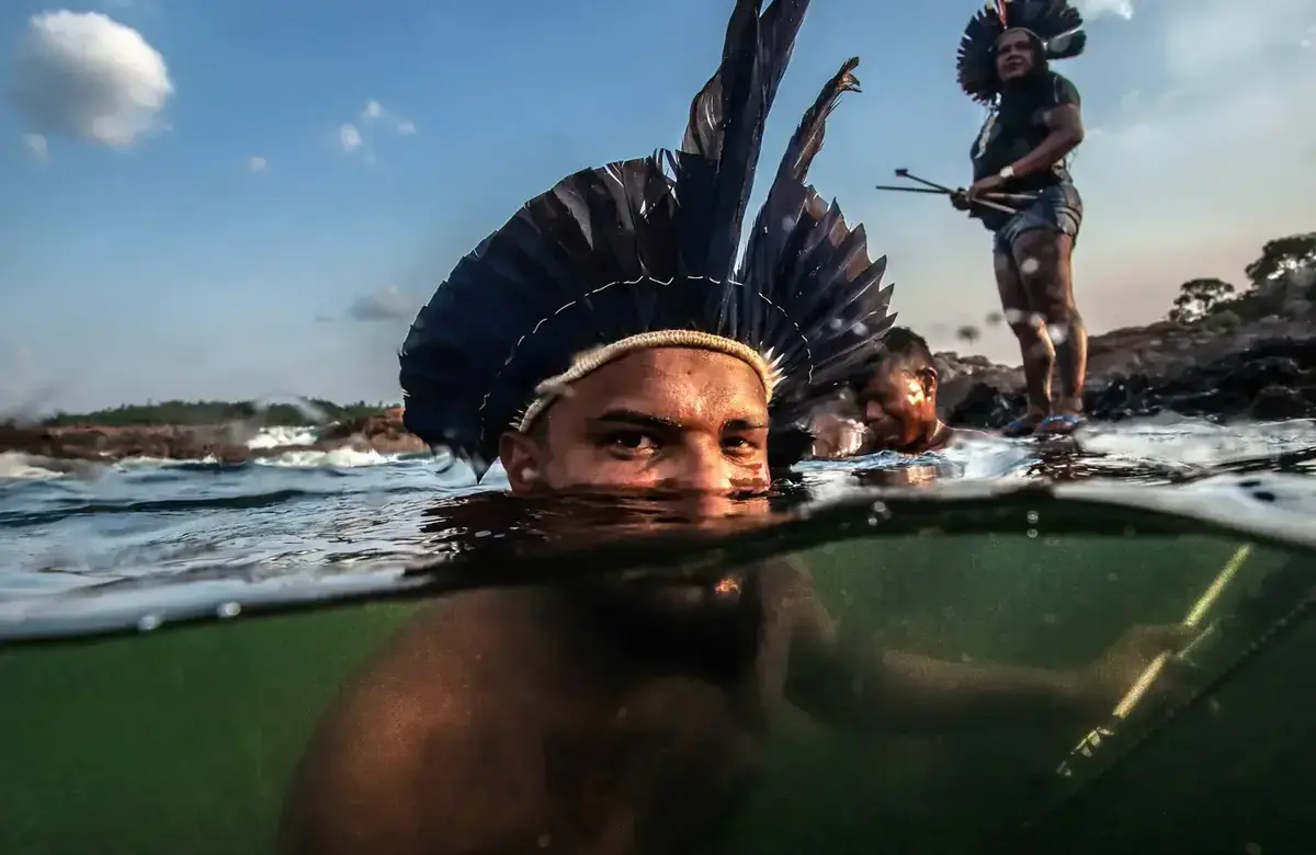 A man with a feathered headdress floats half-submerged in a body of water