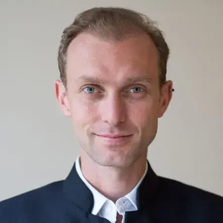 A man wearing a white shirt and dark blue jacket. He has an ear piercing.