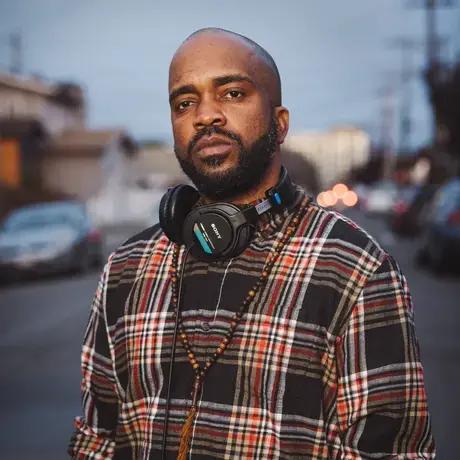 Headshot of Pendarvis Harshaw. He wears a plaid shirt and has headphones around his neck.