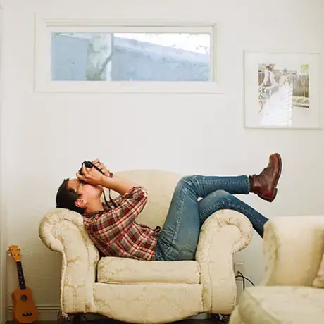 A man lying longways on a chair rests his back against one arm and drapes his legs over the other. He holds a camera to his face.