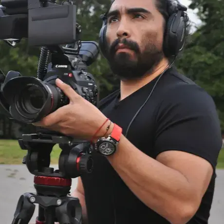Enrique Huaiquil holds a camera that is set up on a tripod. He wears headphones and focuses on the camera.