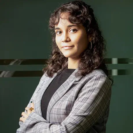 A woman with long curly hair stands against a dark background, arms crossed