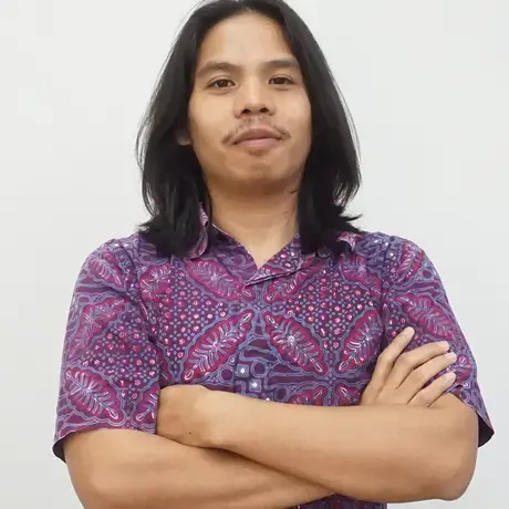 A man with long dark hair and wearing a purple shirt folds his arms