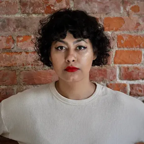 A woman with short curly hair wearing a white shirt stands against a brick wall
