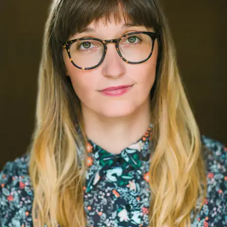 A woman with long hair, bangs, and glasses wears a blue patterned shirt