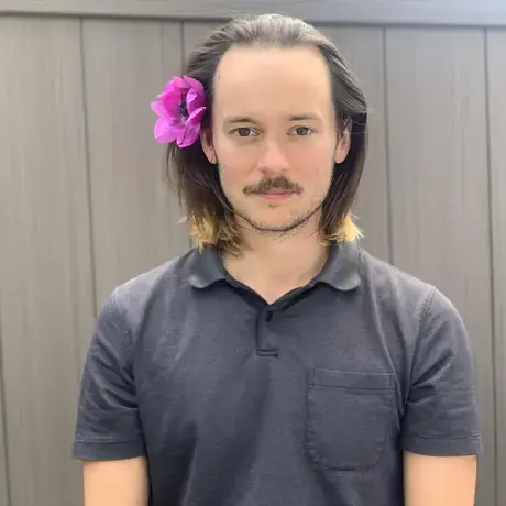 A man with longer brown hair wears a pink flower in his hair and a grey collared shirt