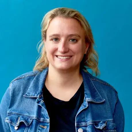 A blonde woman wears a jean jacket against a blue background
