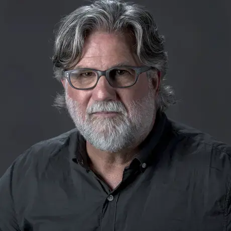 A man with grey hair, a beard, and glasses wearing a grey button-down