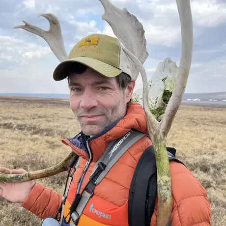 A man beside a large antler bone wearing an orange coat and green baseball cap