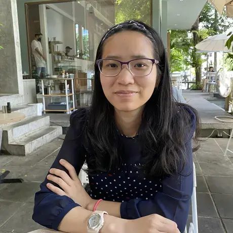 A woman with dark hair and glasses sits at a table with her arms crossed